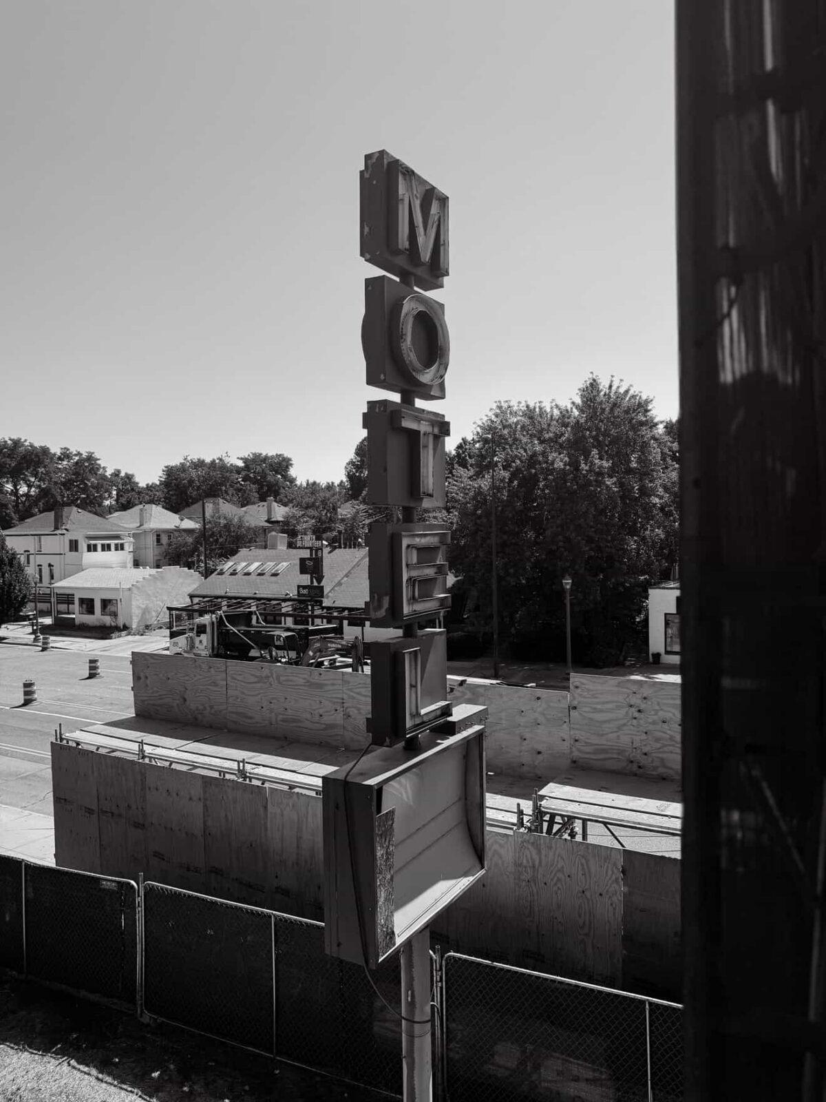 Black-and-white photo of a vertical “MOTEL” sign above a fenced construction area