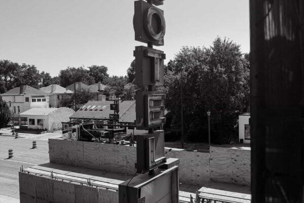 Black-and-white photo of a vertical “MOTEL” sign above a fenced construction area