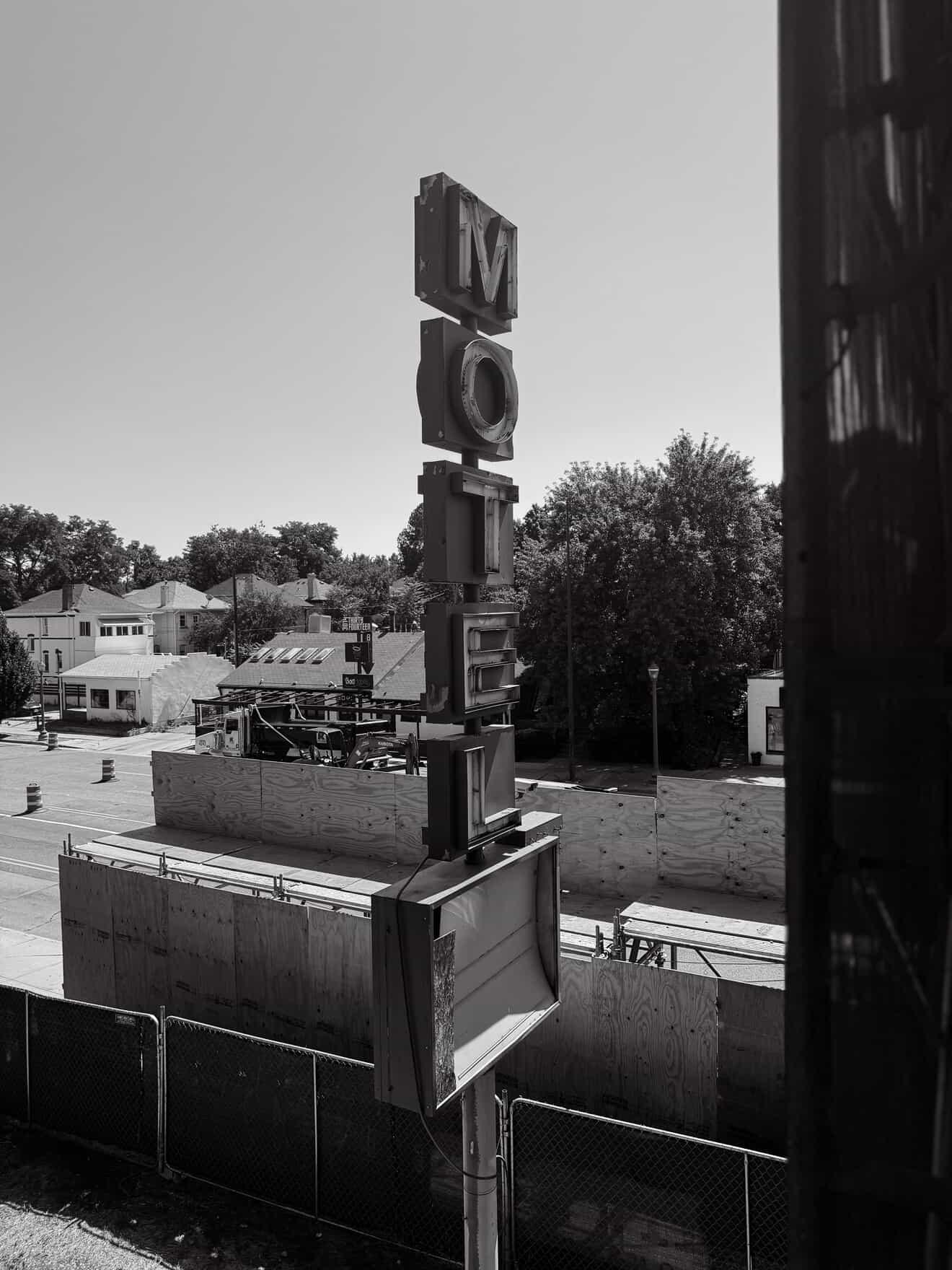 Black-and-white photo of a vertical “MOTEL” sign above a fenced construction area