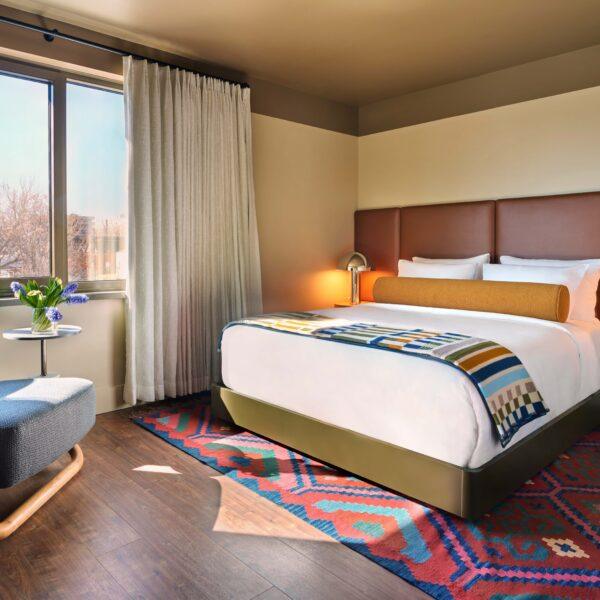Bright hotel bedroom with king bed, patterned throw, red area rug, and sitting chair by a large window