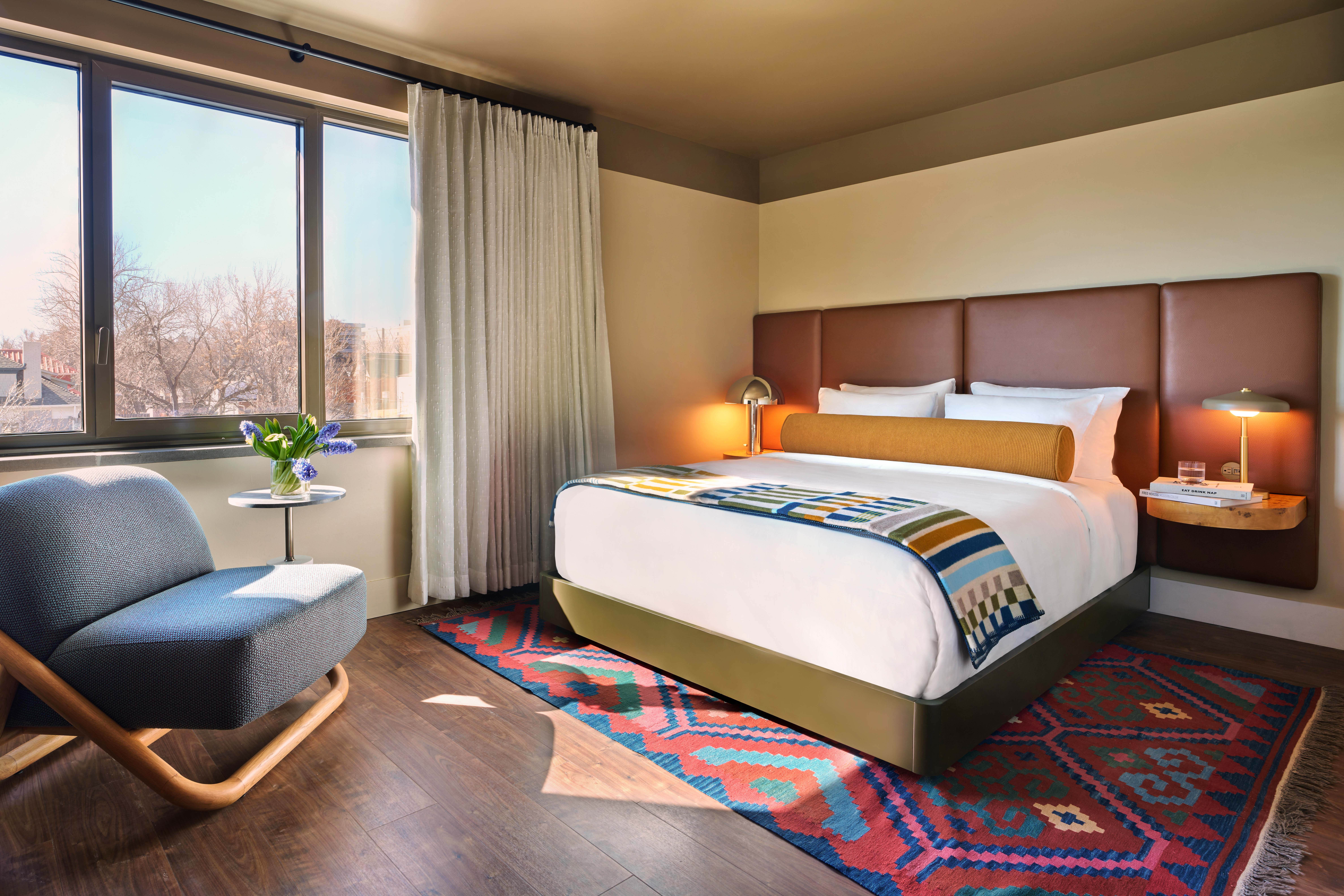 Bright hotel bedroom with king bed, patterned throw, red area rug, and sitting chair by a large window